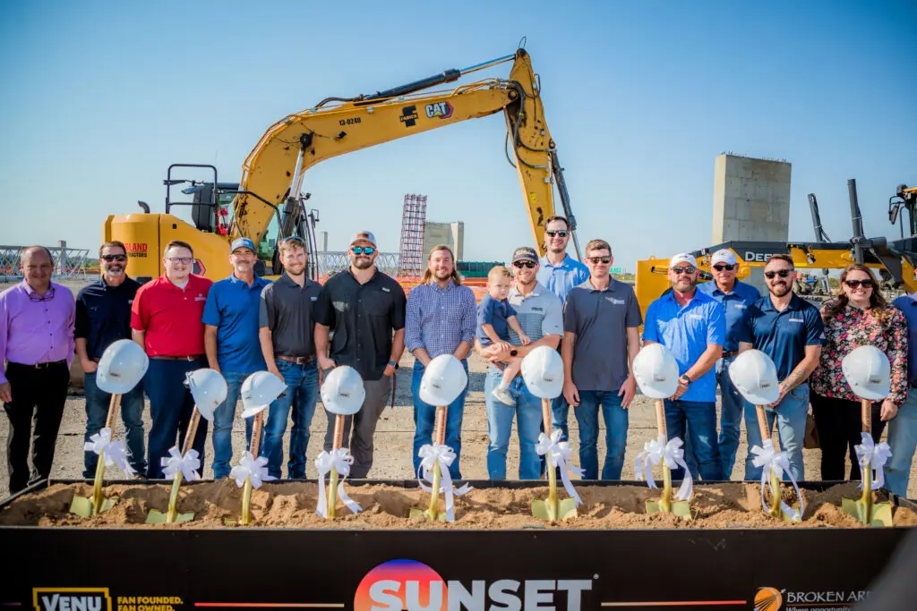A group of people stands in front of construction equipment at a groundbreaking ceremony, with shovels in decorated hard hats lined up in the foreground and a clear blue sky overhead.