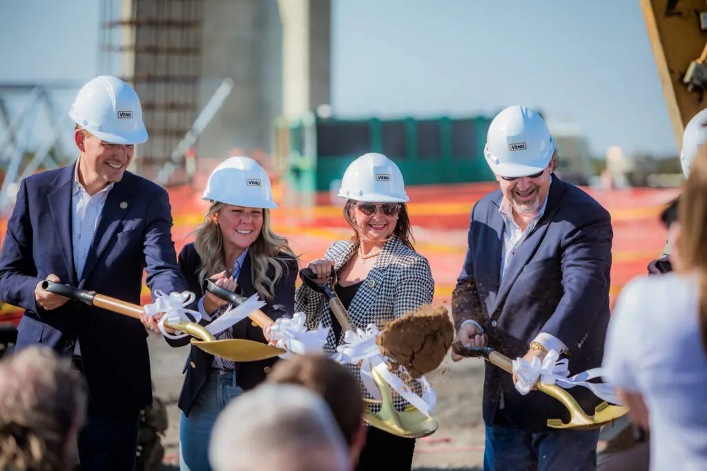 Four people wearing hard hats and business attire smile while holding shovels with white ribbons, participating in a groundbreaking ceremony at a construction site. Dirt is being turned over, and construction equipment is visible in the background.