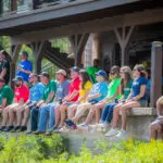 A group of people wearing colorful Crossland-branded t-shirts sit on a ledge outdoors, attentively watching an event. The participants appear engaged, with some wearing sunglasses and holding drinks. The background features a wooden deck with additional spectators standing and a staircase leading down to the gathering area. The setting is lush with greenery, indicating a relaxed, outdoor event.