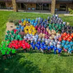 A large group of people stands on grass, organized by shirt color into several groups, including green, red, yellow, blue, orange, maroon, gray, black, and white, in front of a building.