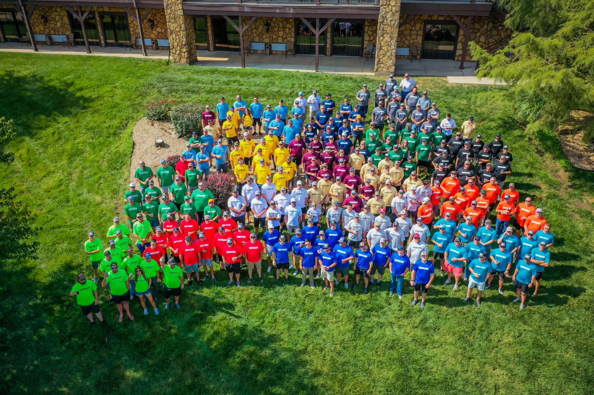 A large group of people stands on grass, organized by shirt color into several groups, including green, red, yellow, blue, orange, maroon, gray, black, and white, in front of a building.