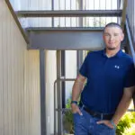 A man wearing a navy blue polo shirt and jeans leans casually against a metal railing under an outdoor staircase, smiling at the camera. Sunlight illuminates the scene and there is greenery in the background.