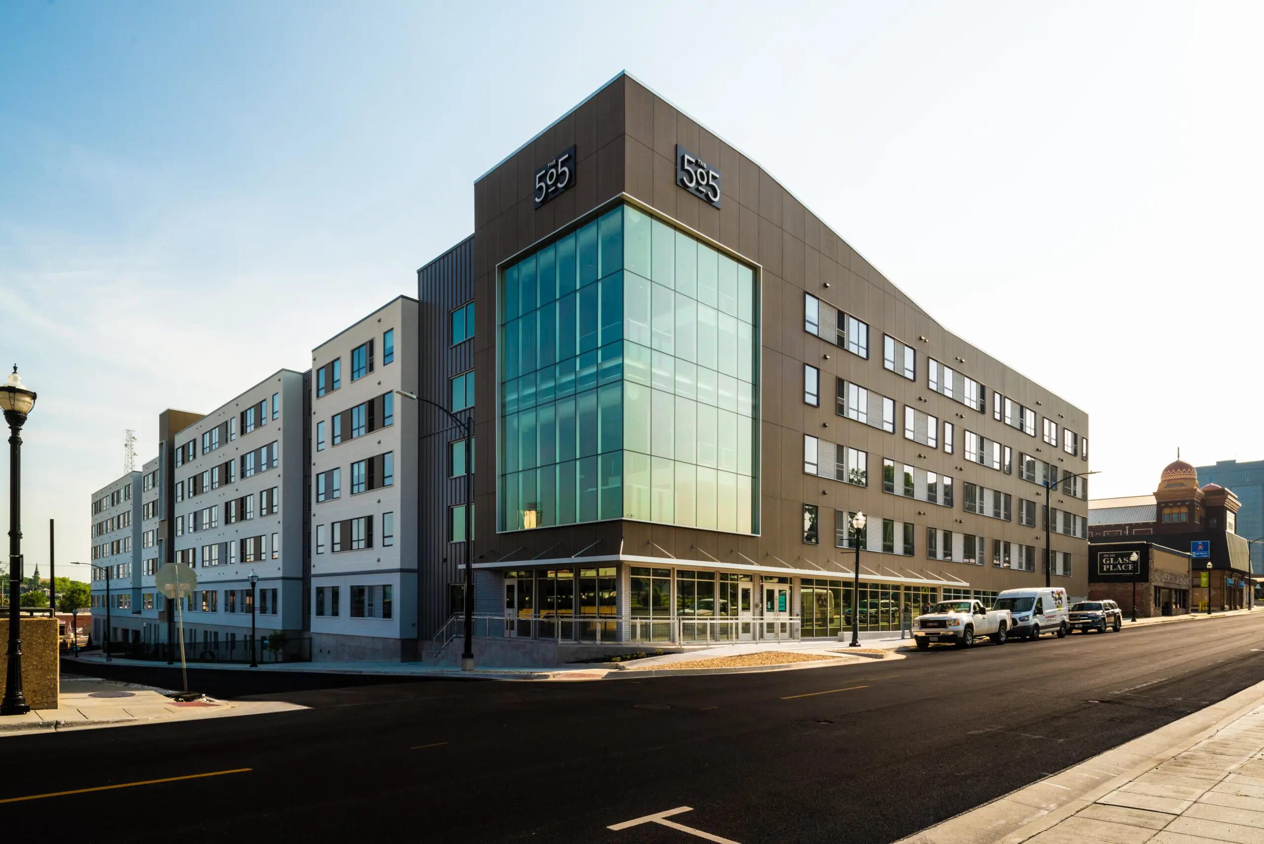 A modern, multi-story apartment building with large glass windows on the corner and “555” displayed on its facade, located on a clean, empty street with a few parked cars and clear sky above.