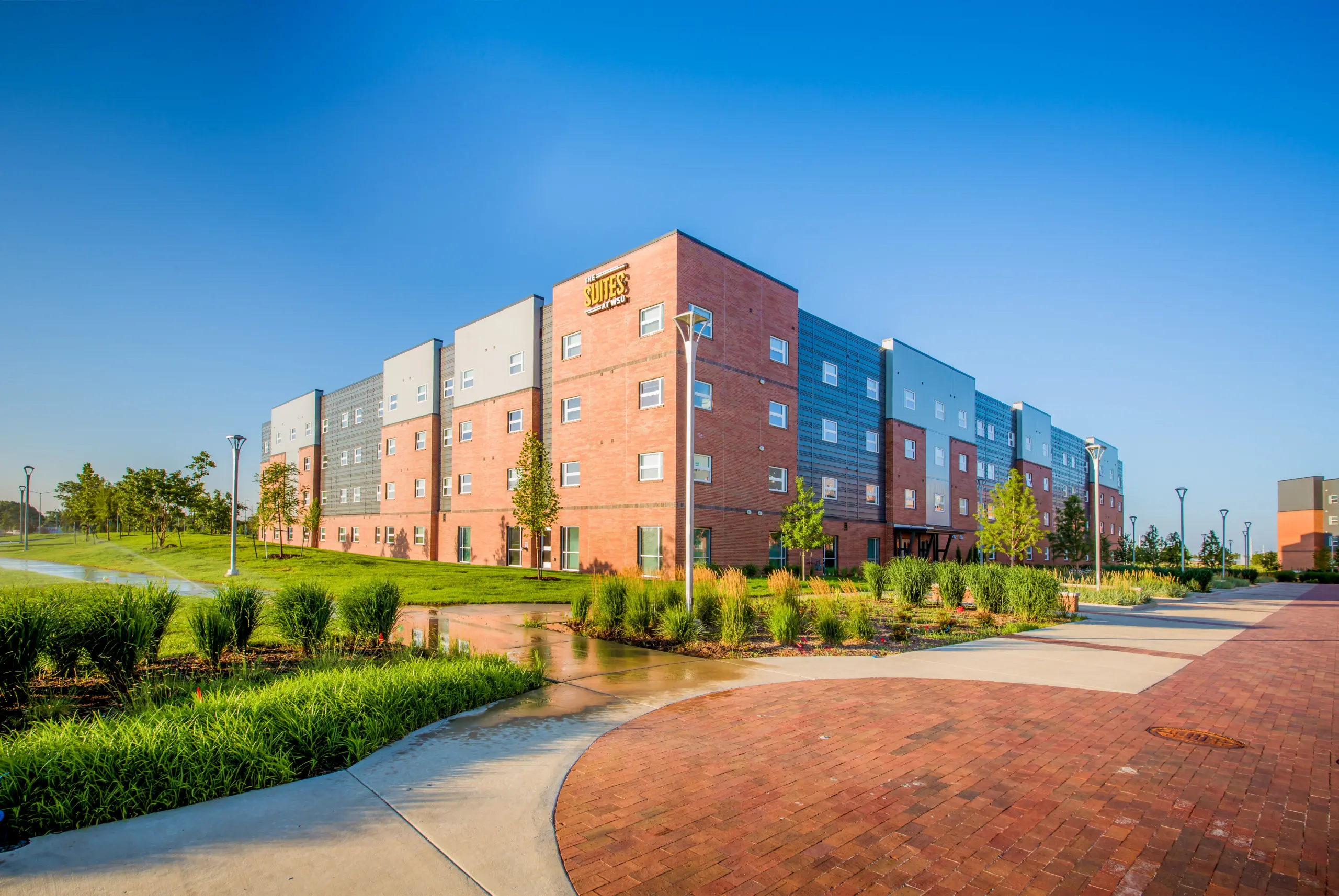 Modern multi-story brick and gray building labeled “Suites,” surrounded by green lawns, landscaped plants, trees, and paths under a clear blue sky. Sprinklers water the grass in the foreground.