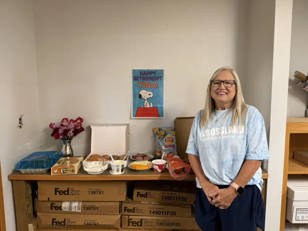A smiling woman with long gray hair and glasses stands by a table with snacks and treats. Behind her is a "Happy Retirement Tina!" sign with Snoopy. The table sits on stacked FedEx boxes in an office setting.