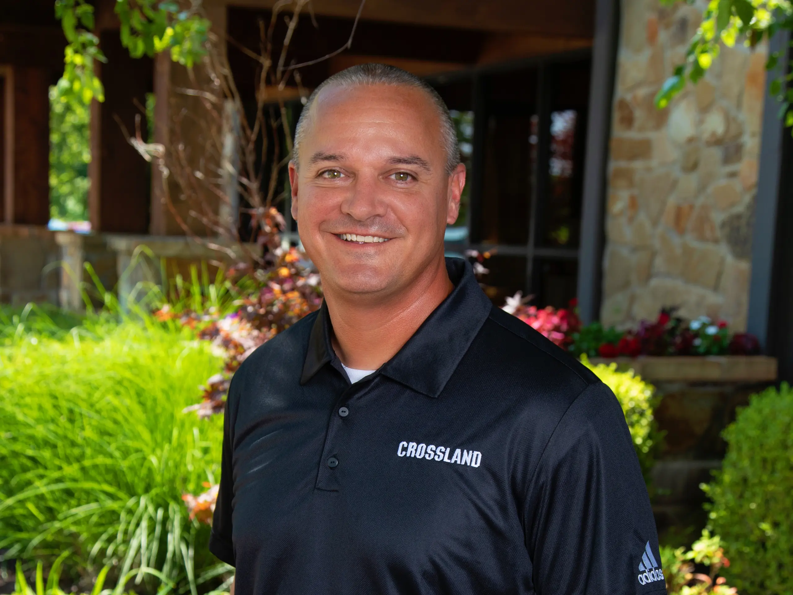 A man wearing a black "CROSSLAND" polo shirt stands outside in front of a stone building with green plants and flowers, smiling at the camera.