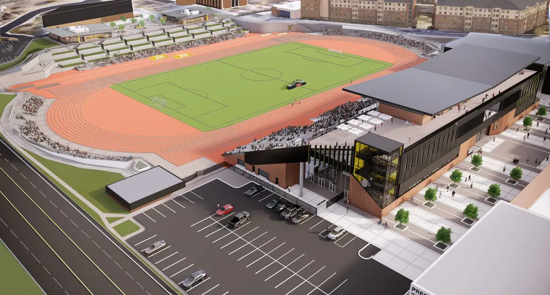 Aerial view of a modern sports stadium with a soccer field surrounded by a running track, stands filled with people, adjacent parking lot, and nearby buildings in the background.