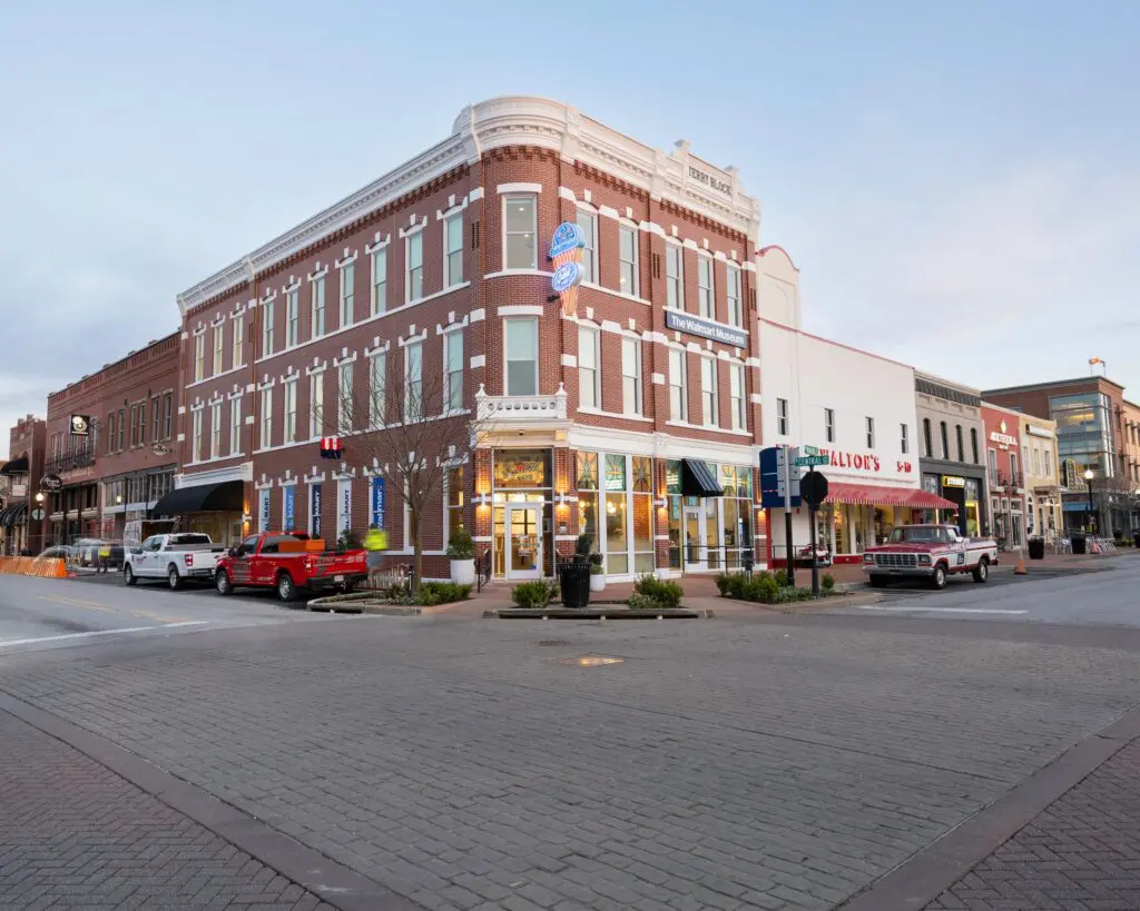 A red brick corner building with white trim houses shops and restaurants on a quiet street, with several parked trucks and a few small trees along the sidewalk. The sky is clear and buildings line both sides of the street.