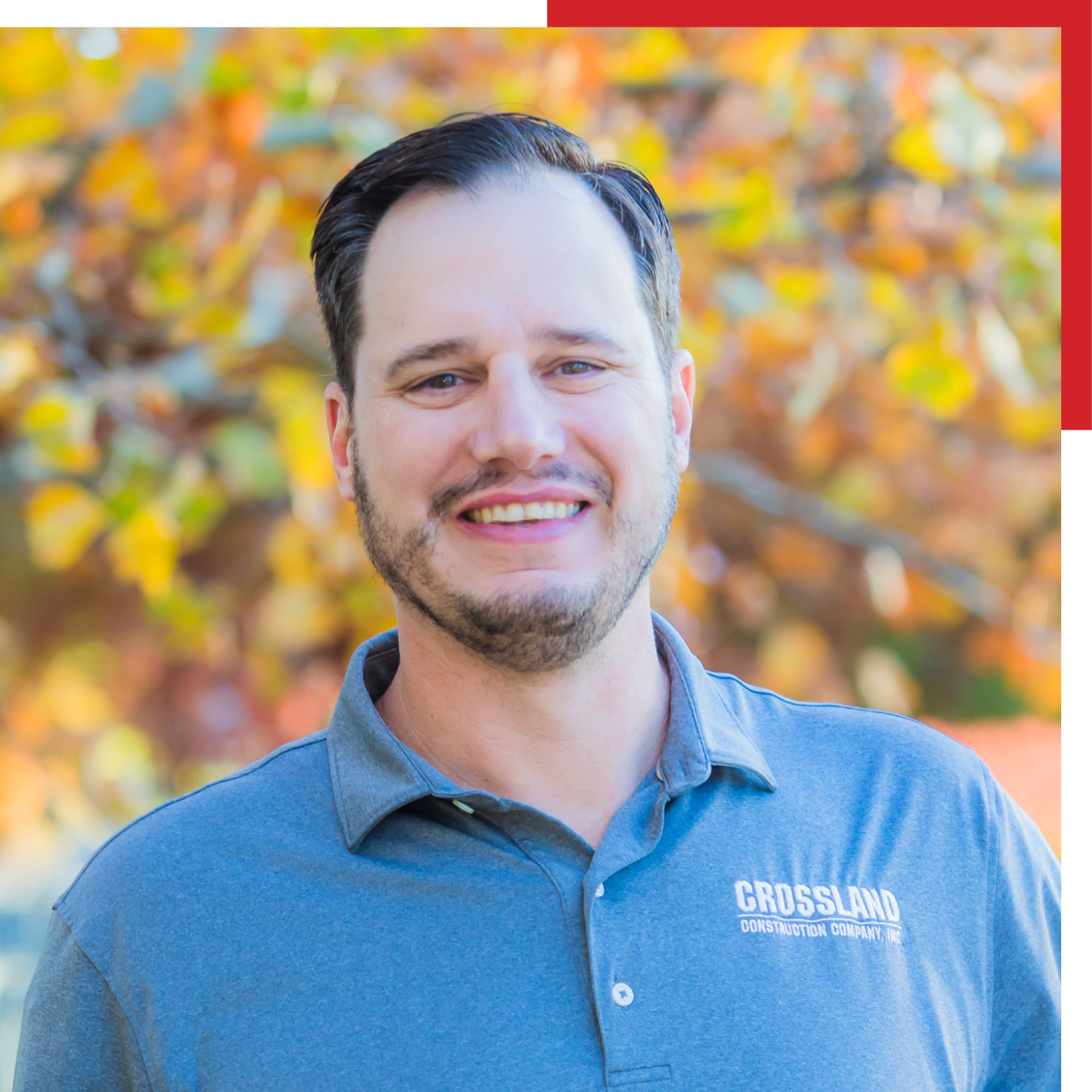 A man with dark hair and a trimmed beard smiles outdoors, wearing a gray "Crossland Construction Company, Inc." polo shirt. The background features colorful autumn leaves out of focus.