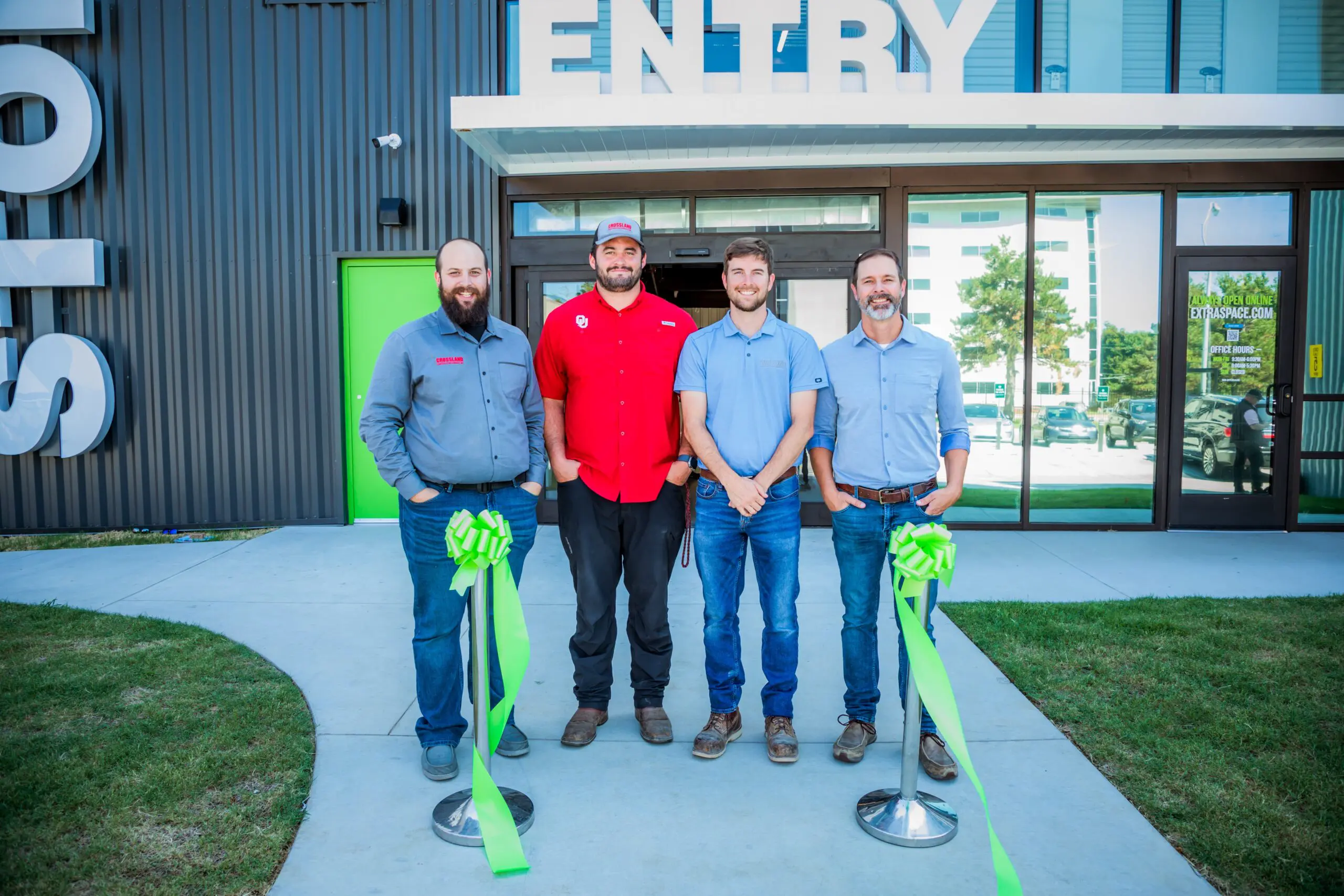 Four men stand smiling in front of a building entrance, flanked by two green ribbon bows on stands. The group appears to be at a ribbon-cutting event. The entrance sign and modern building glass are visible behind them.