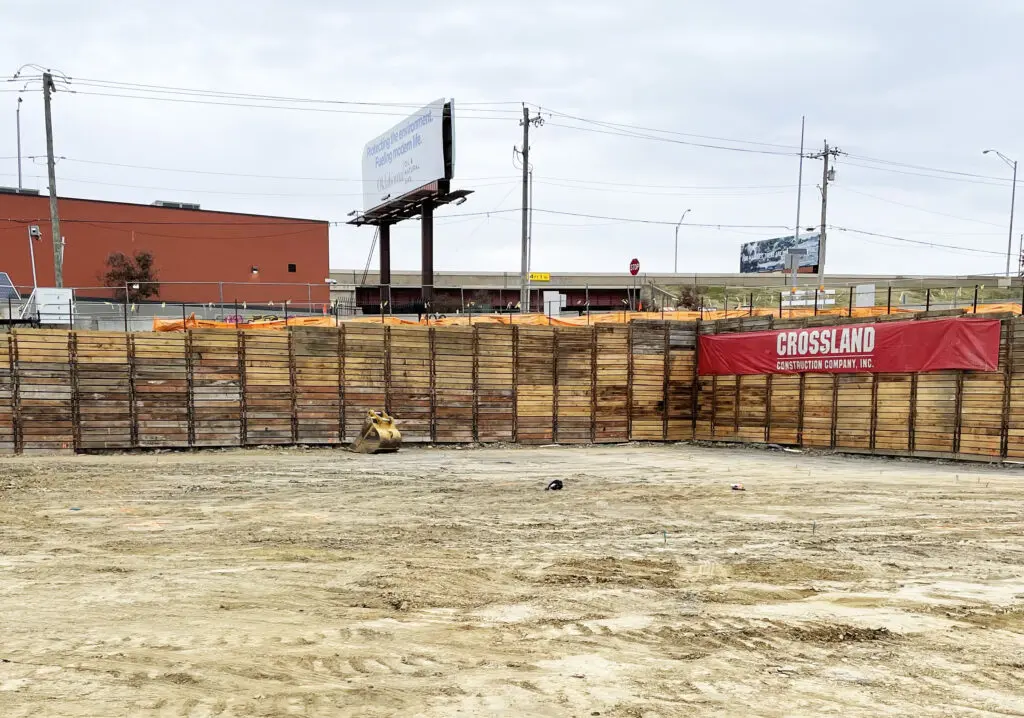 A construction site with a dirt ground, wooden retaining walls, a red banner reading "CROSSLAND CONSTRUCTION COMPANY, INC.," and an orange safety barrier. A billboard and street are visible in the background.