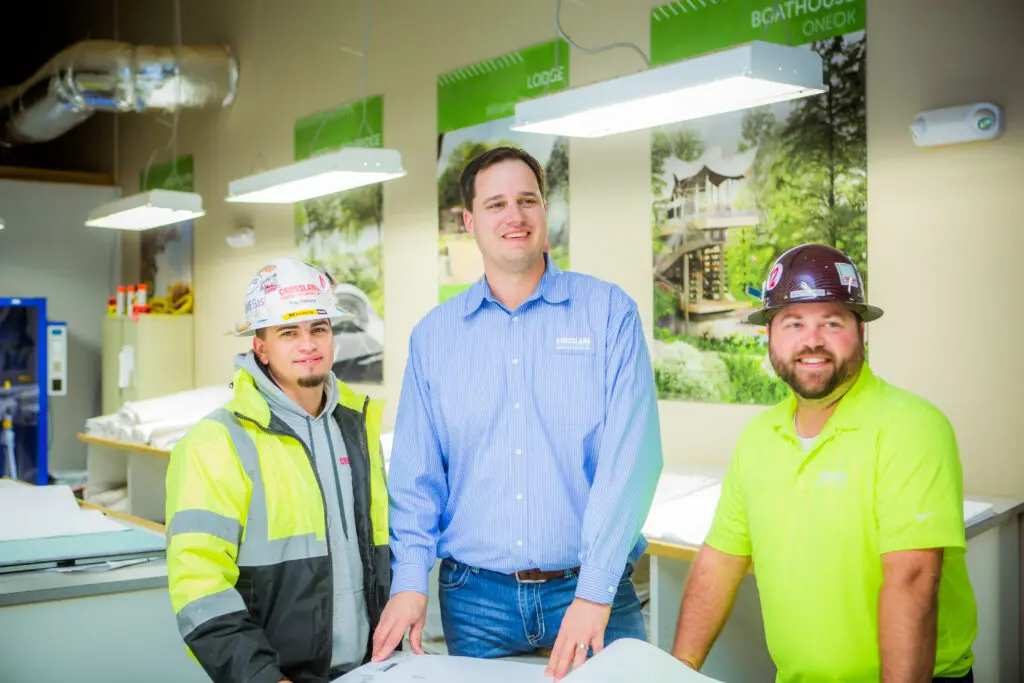 Three men, two in construction gear and one in business attire, stand smiling around a table with blueprints in a brightly lit office featuring nature-themed posters on the walls.