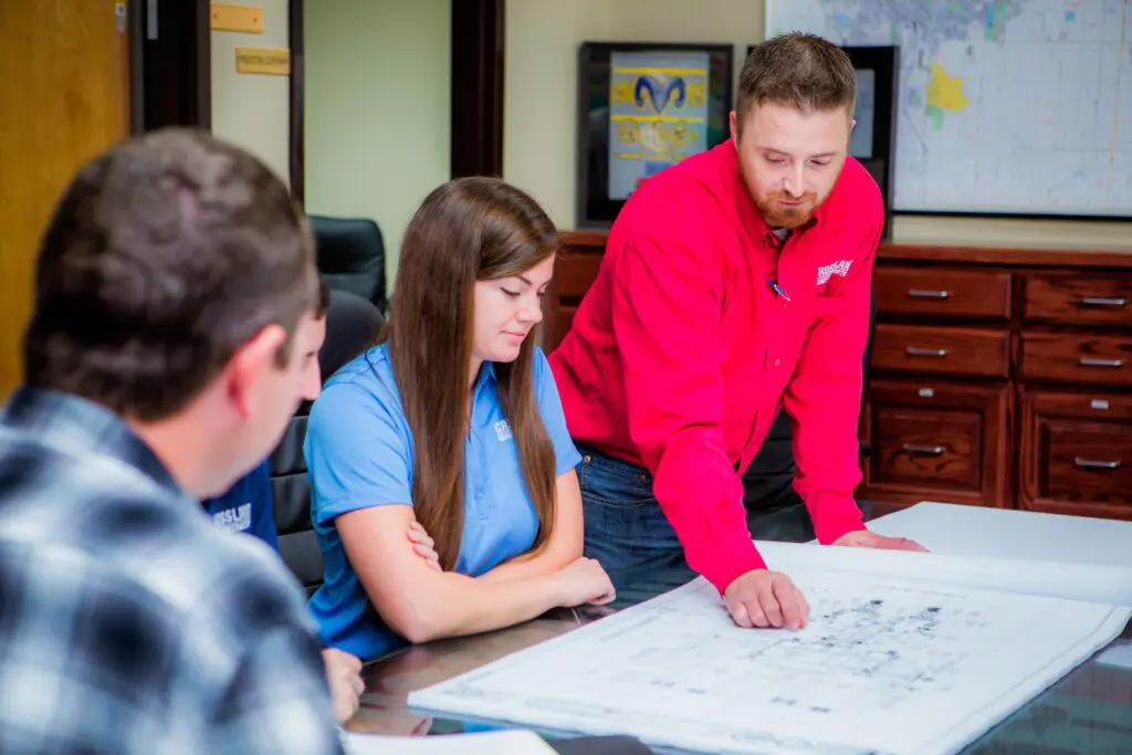 A man in a red shirt points to a large blueprint on a table while three colleagues listen and look on in a meeting room with maps and filing cabinets in the background.
