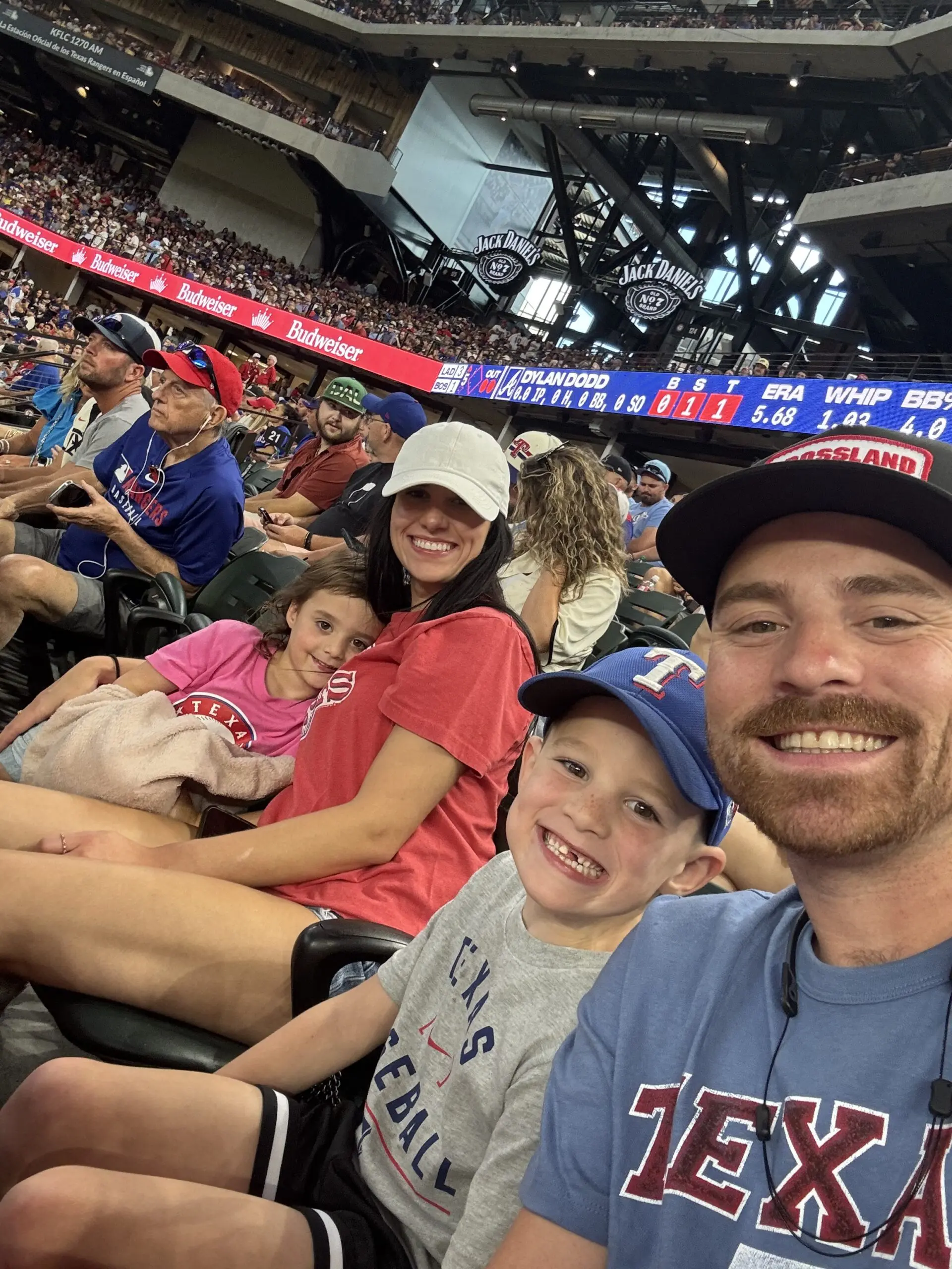 A smiling family of four wearing Texas Rangers gear sits in stadium seats at a baseball game, surrounded by other fans. The stands are full and the field is visible in the background.