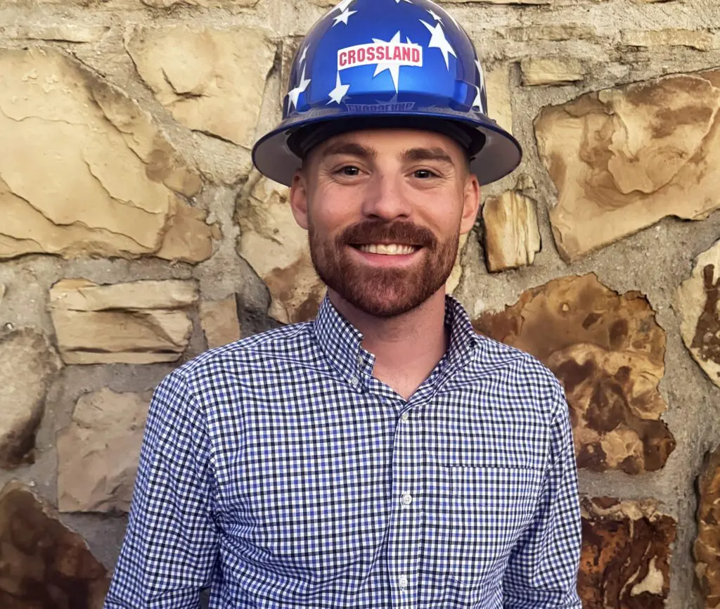 A smiling man with a reddish beard, wearing a blue hard hat with white stars and a "CROSSLAND" sticker, stands in front of a stone wall. He is dressed in a checked button-up shirt.