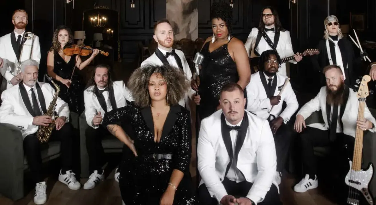 A group of thirteen musicians dressed in black and white formal attire pose together indoors, some holding instruments like a violin, saxophone, and bass guitar, with elegant dark décor in the background.