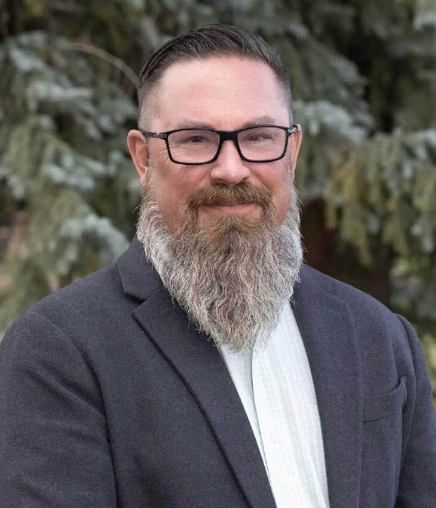 A man with a long gray beard, glasses, and short dark hair, wearing a dark blazer and light shirt, stands outdoors in front of green trees, smiling slightly at the camera.