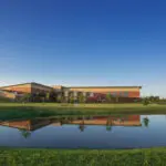 A modern, low-rise building with tan and brick exterior is reflected in a small pond, surrounded by green grass and trees under a clear blue sky.