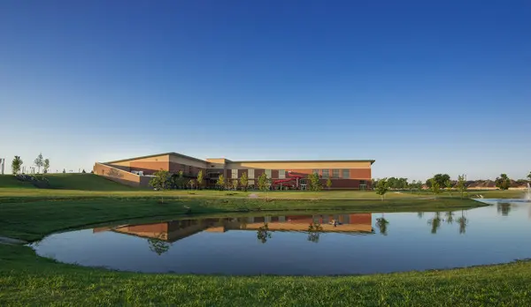 A modern, low-rise building with tan and brick exterior is reflected in a small pond, surrounded by green grass and trees under a clear blue sky.