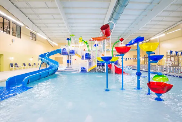 An indoor aquatic play area with shallow water, colorful slides, large water buckets, and various interactive water features under a high ceiling with exposed ducts. Blue chairs line the walls in the background.