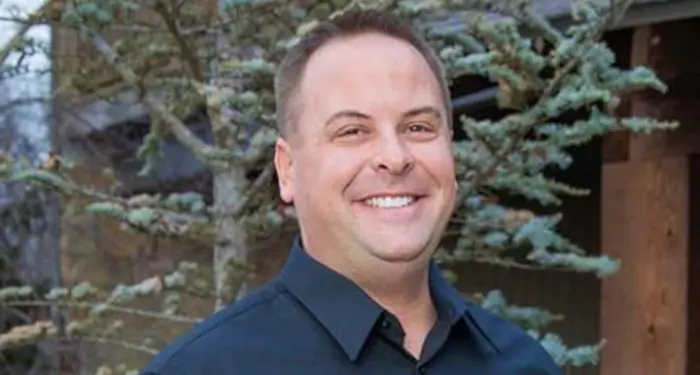 A man with short light brown hair, wearing a dark button-up shirt, smiles while standing outdoors in front of green trees and a wooden building.