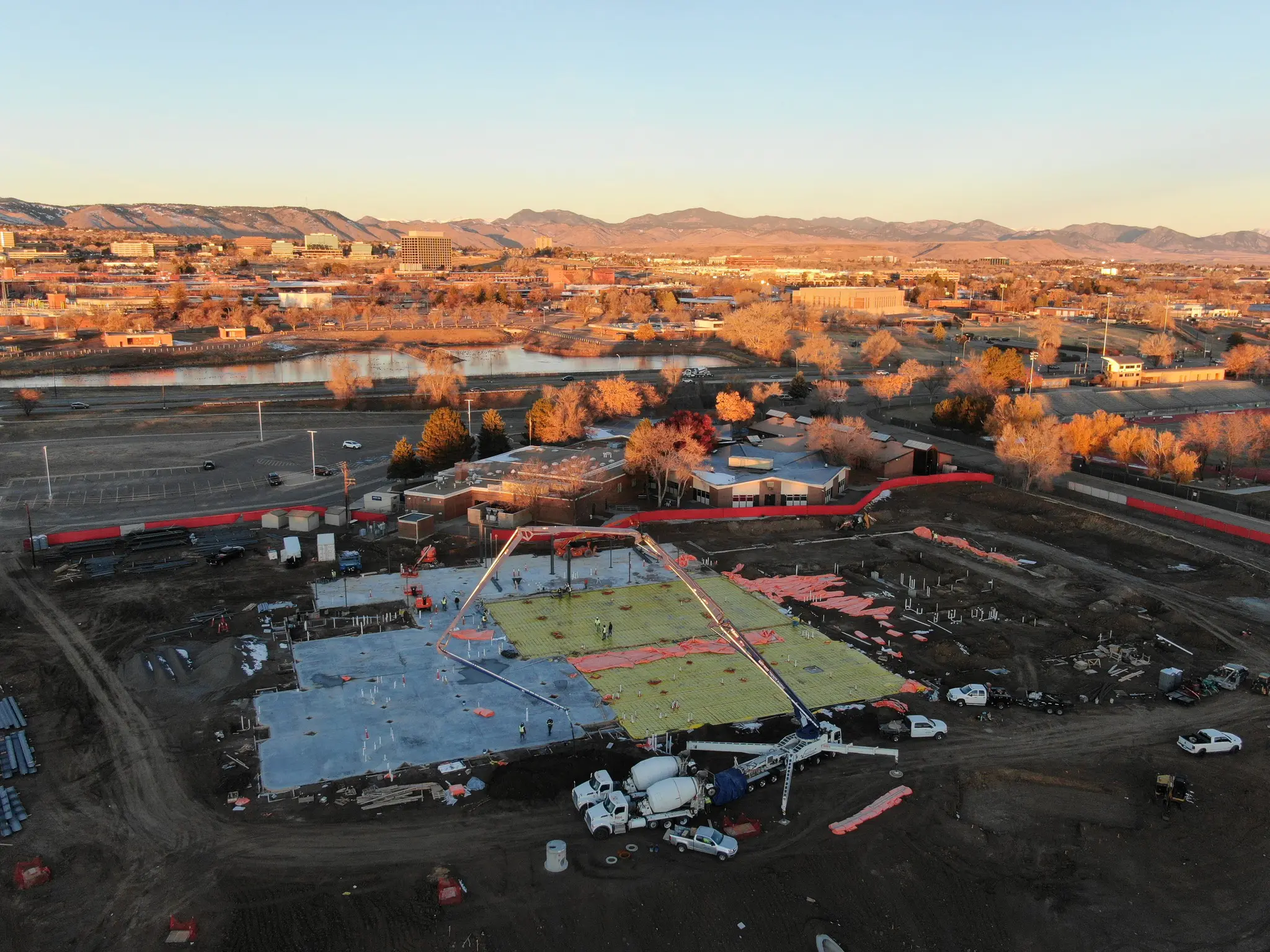 Aerial view of a construction site with concrete foundation work in progress, surrounded by vehicles and equipment, with a town and mountains visible in the background under a golden sunset.
