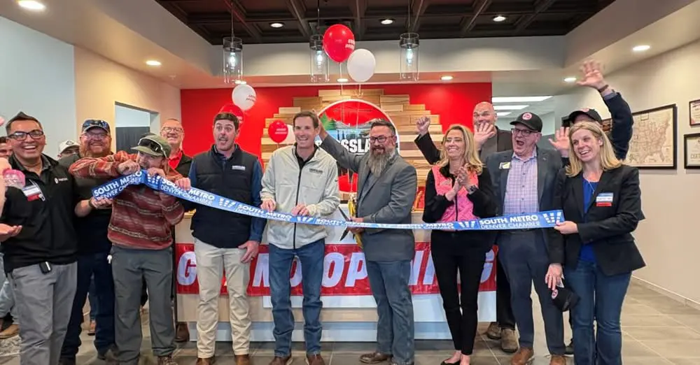 A group of people smile and cheer while holding a blue South Metro Chamber ribbon at a business ribbon-cutting ceremony indoors, with balloons and a red wall in the background.
