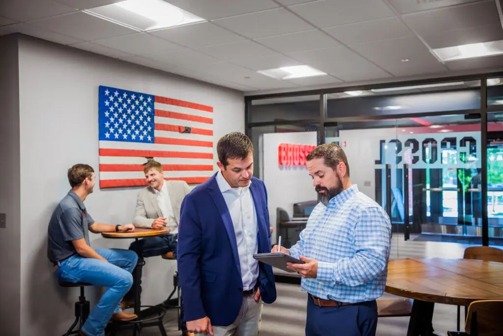 Two men in business attire discuss something on a tablet in an office lounge. In the background, two others sit and talk near a wall with an American flag decoration.