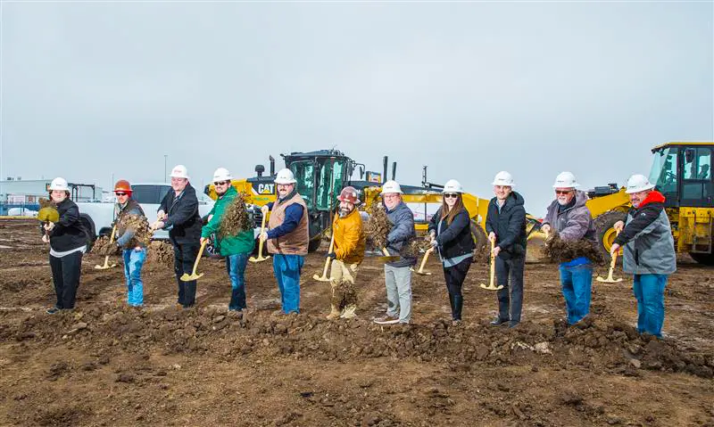 A group of people wearing hard hats and construction gear stand in a line, holding shovels and digging into the dirt at a construction site, with heavy machinery visible in the background.