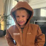 A young boy smiles indoors while wearing a brown hooded jacket with "CROSSLAND" and "Cam" embroidered on it. He stands in front of a fireplace, and sunlight streams through the window behind him.