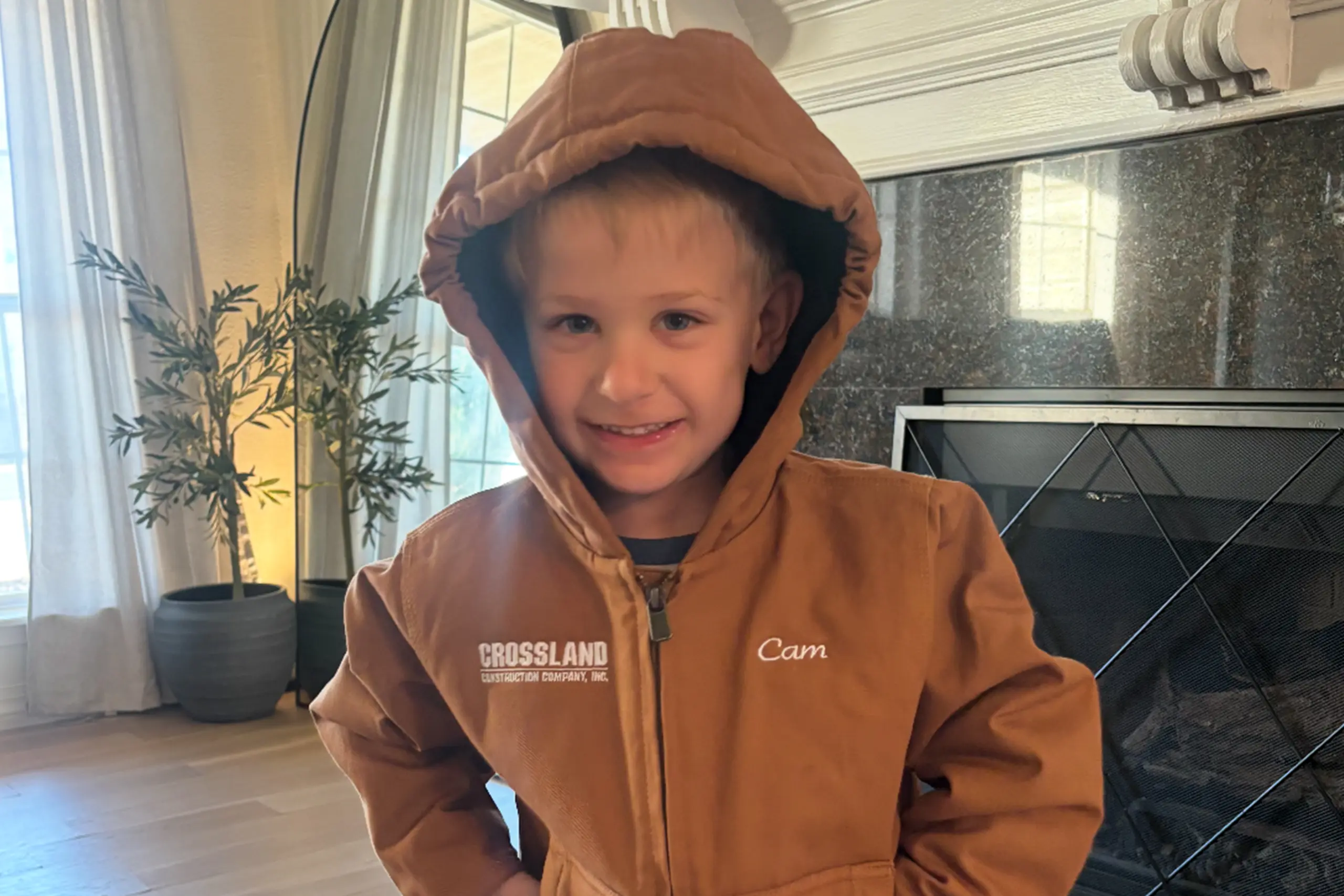 A young boy smiles indoors while wearing a brown hooded jacket with "CROSSLAND" and "Cam" embroidered on it. He stands in front of a fireplace, and sunlight streams through the window behind him.