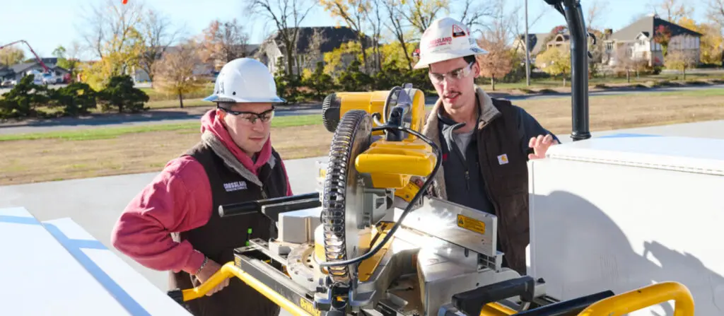 Two construction workers wearing hard hats and safety glasses operate and inspect a yellow power saw outdoors on a sunny day, with trees and houses visible in the background.