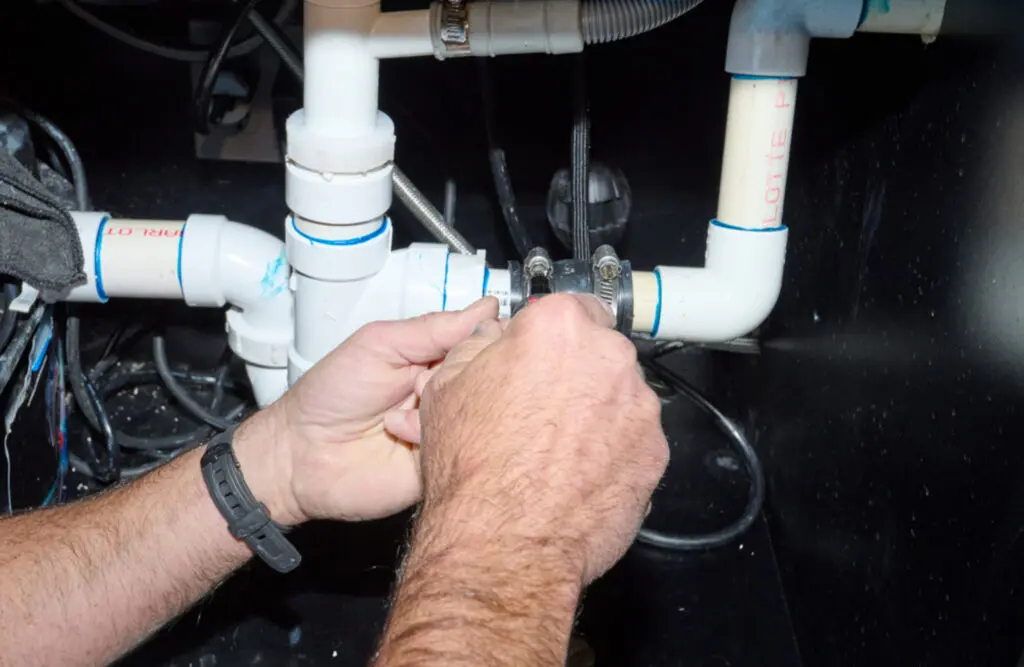 A person’s hands repairing white PVC pipes under a sink, tightening connections with a wrench. Black cables and other plumbing components are also visible in the background.