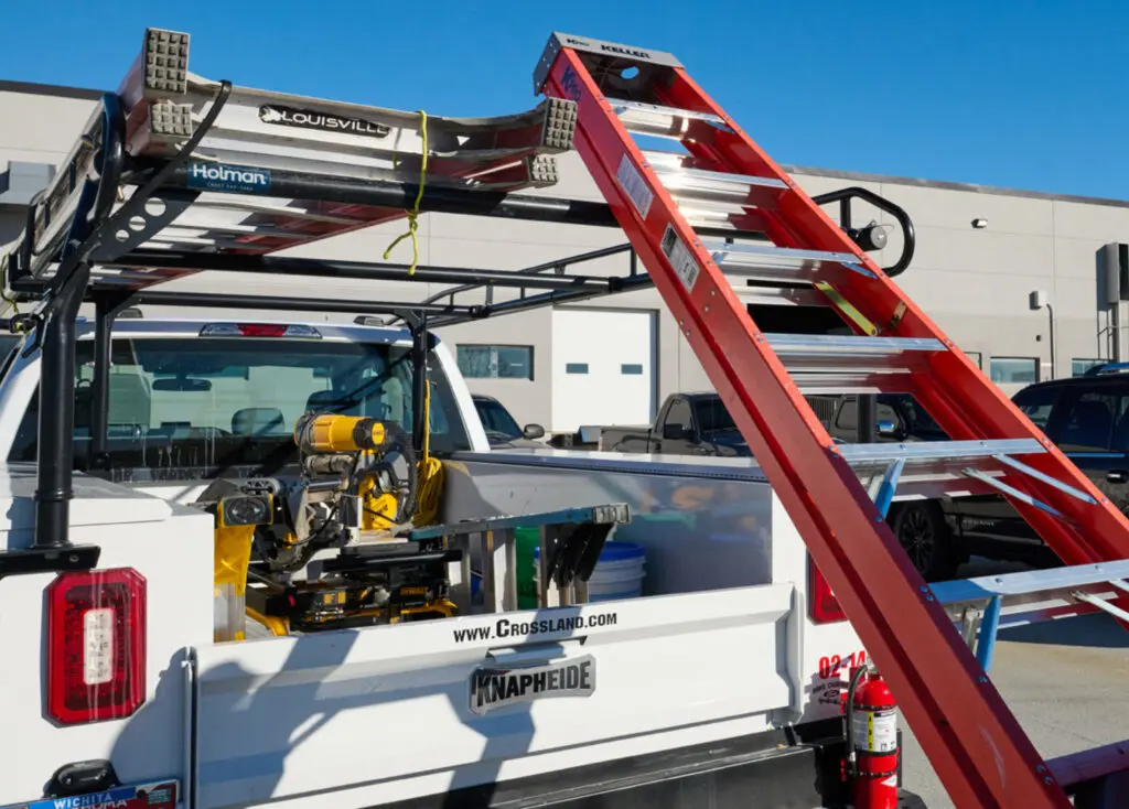 A white pickup truck with a mounted tool rack holds ladders, a saw, and construction tools. A large red ladder leans into the truck bed. Buildings and parked vehicles are visible in the background on a sunny day.