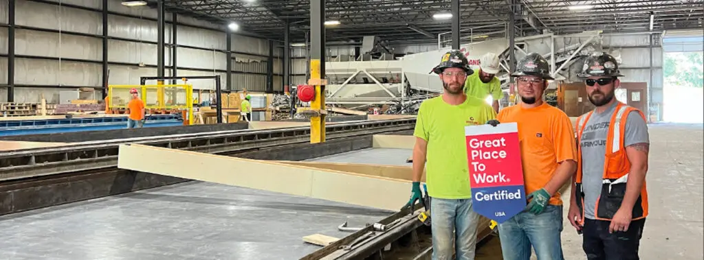 Three Crossland construction workers in safety gear stand inside a large factory, holding a "Great Place To Work Certified" sign. Industrial equipment and building materials are visible in the background.