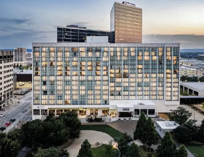 A modern multi-story hotel building with large windows, illuminated rooms, and a landscaped park with trees in the foreground, set against a cityscape at dusk.