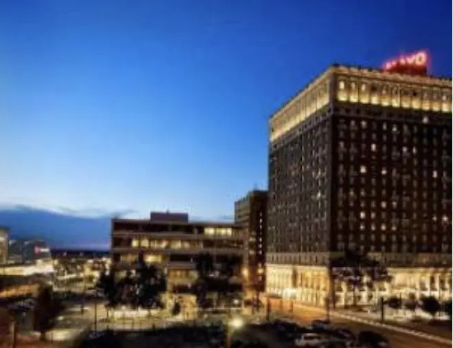 A cityscape at dusk featuring a tall, illuminated historic hotel with a red neon sign on its roof, surrounded by other buildings and streetlights. The sky is a deep blue, and cars are visible on the street below.
