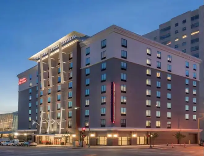 A modern, multi-story hotel building at dusk with large windows and illuminated signs, located on a street corner in an urban area.