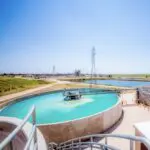 A large circular water treatment basin with turquoise water, surrounded by safety railings and industrial equipment, set in an open landscape under a clear blue sky.