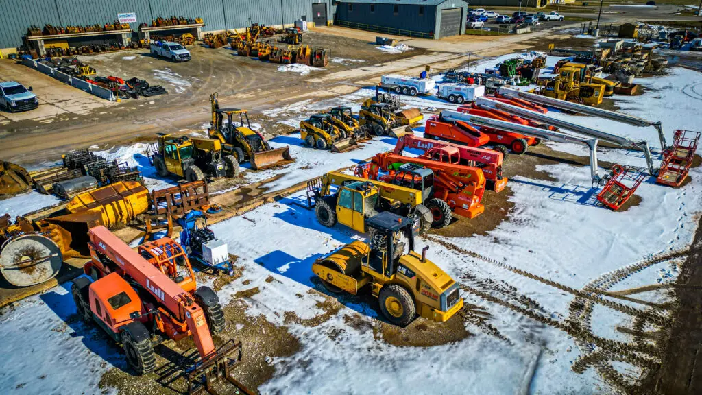 Aerial view of a snow-dusted industrial lot displaying various construction vehicles and heavy machinery, including forklifts, loaders, lifts, and utility trucks, parked in organized rows.