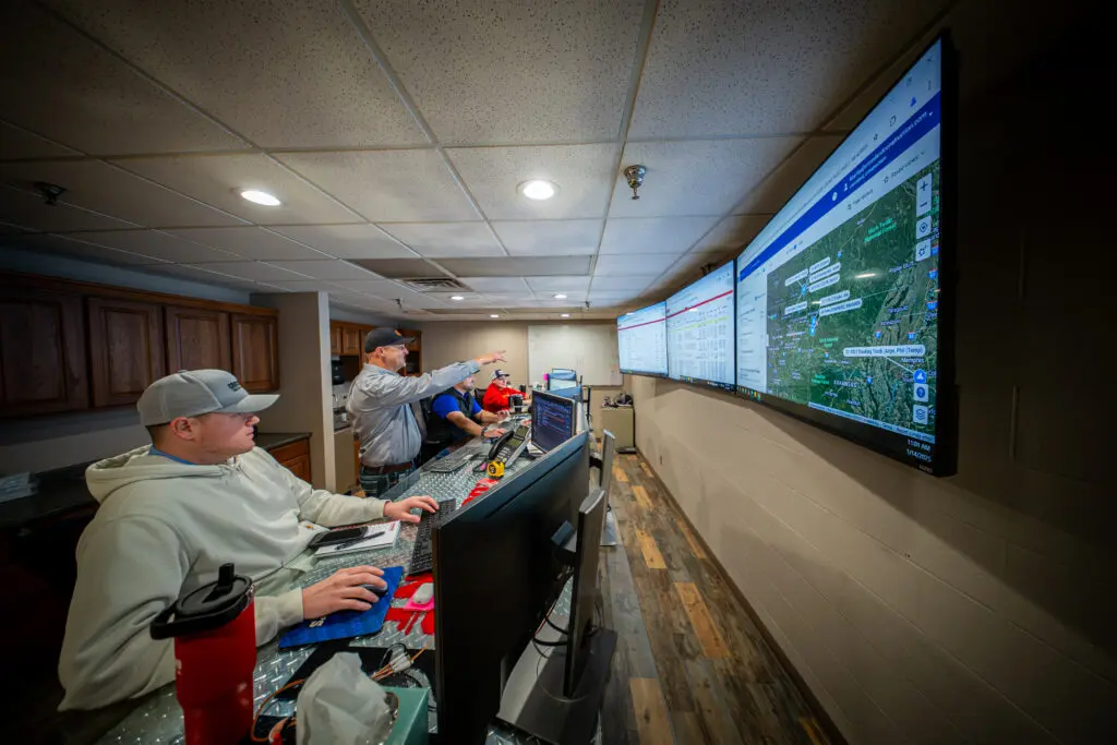 Three people sit at a control room desk with multiple monitors, while one person stands and points at large wall-mounted screens displaying maps and data. The room has wood flooring and a drop ceiling.