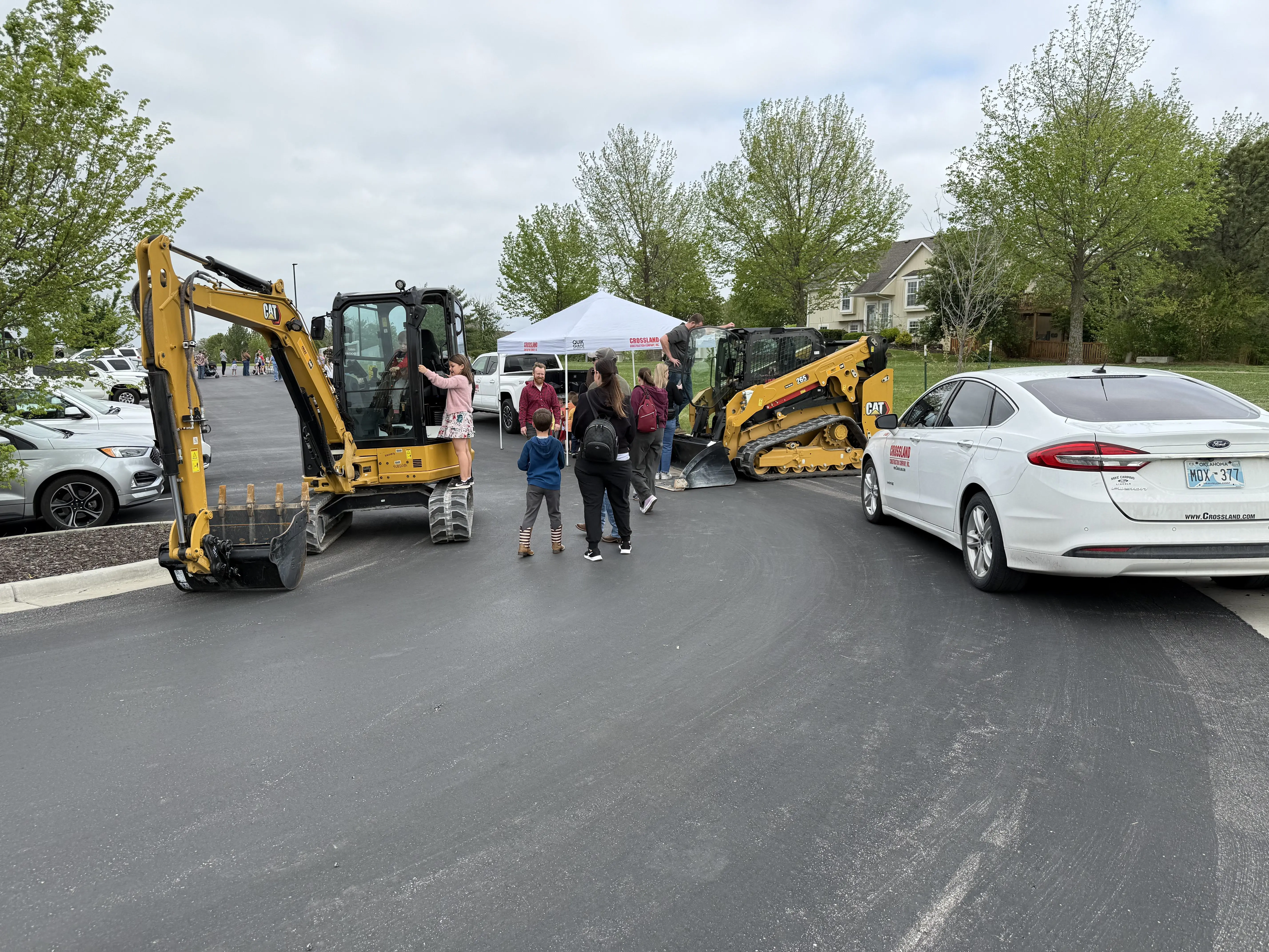 A group of people, including children, gather near two construction vehicles and a white tent in a parking lot; a white car is parked nearby, and trees and houses are visible in the background.
