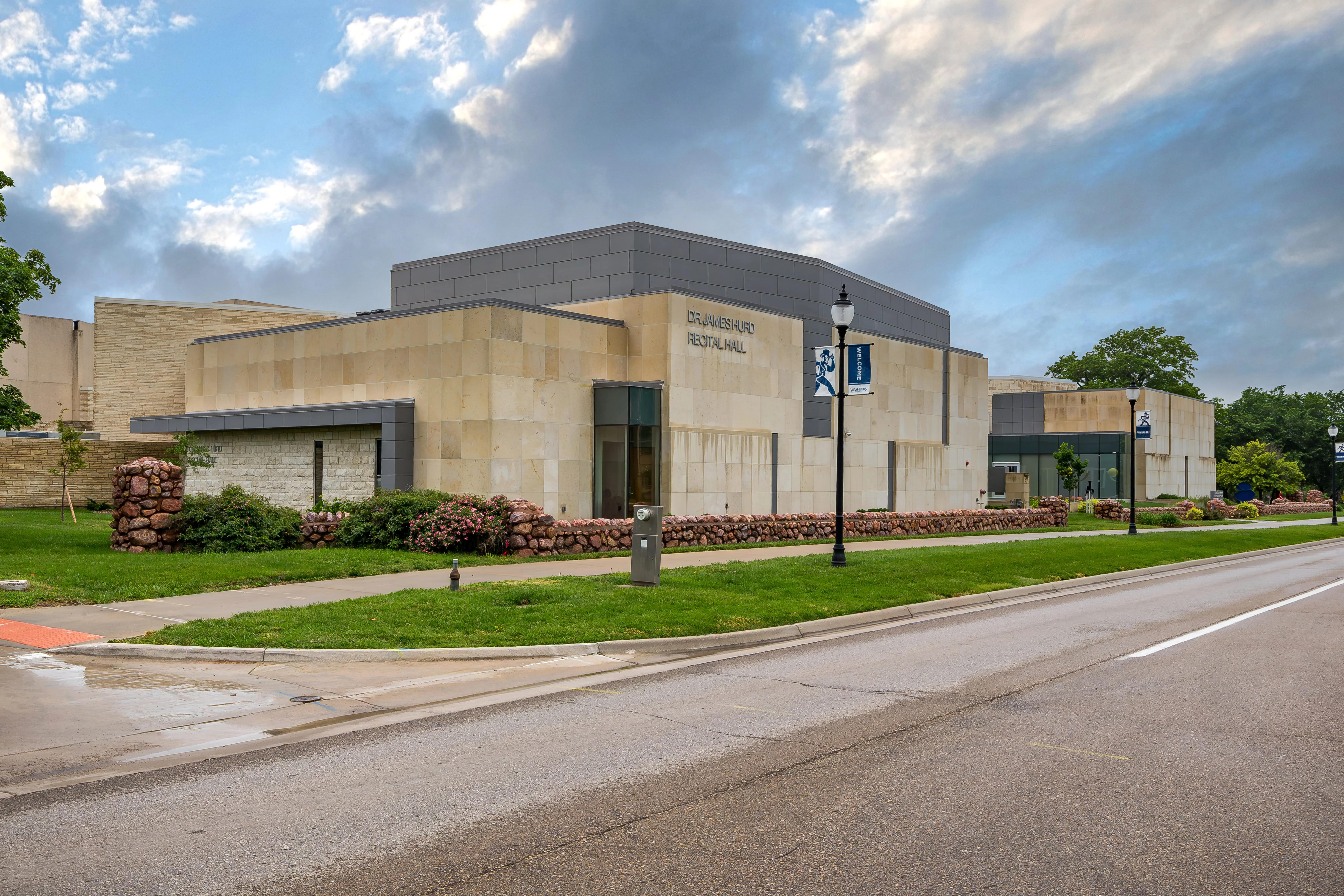 A modern, light stone and gray building labeled "Brannigan Recital Hall" sits beside a street, with green grass, landscaped bushes, and a few trees under a cloudy sky.