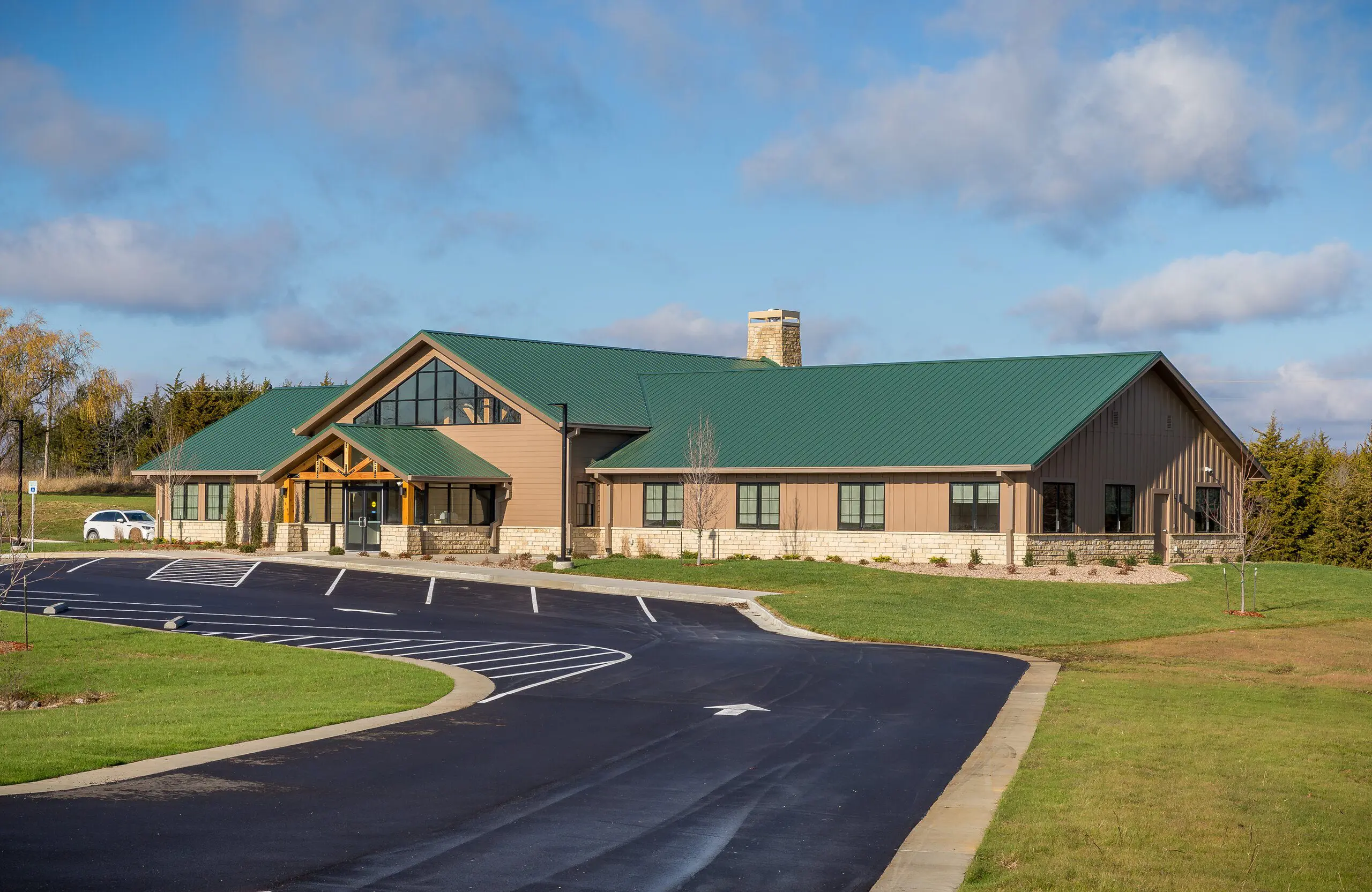 A one-story building with a green metal roof, large windows, and a wooden entrance sits next to an empty parking lot, surrounded by grass and trees under a partly cloudy sky.