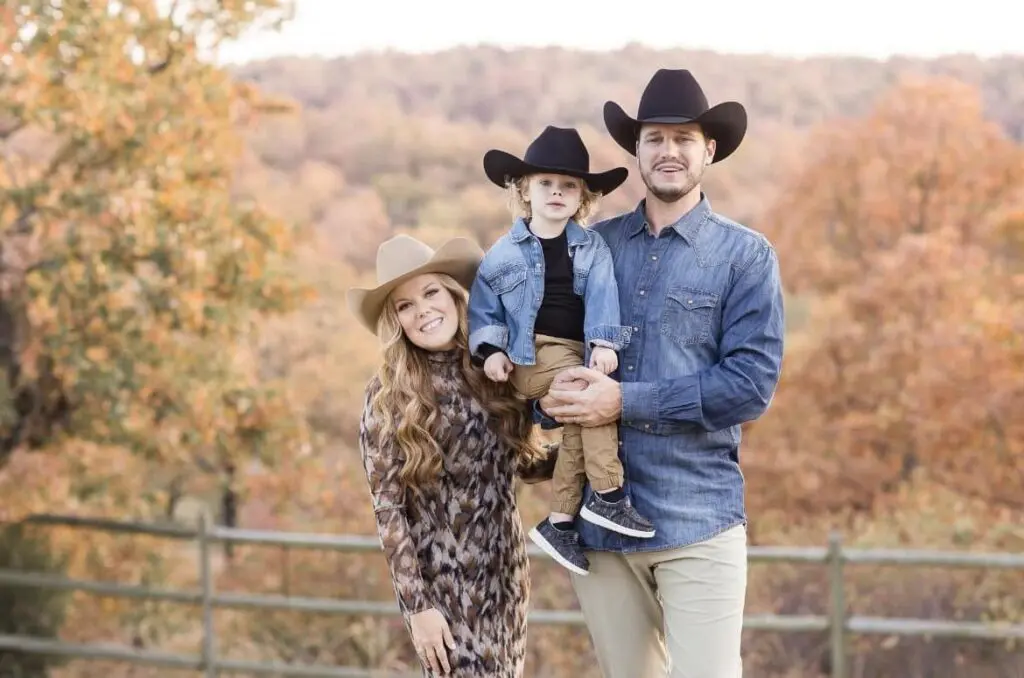 A smiling family of three wearing cowboy hats poses outdoors in front of autumn trees. The man holds a young child, while the woman stands next to them, all dressed in casual fall outfits.