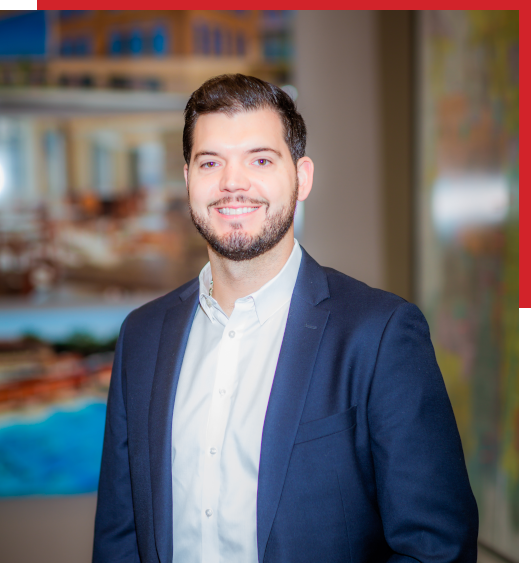 A man with short dark hair and a beard, wearing a navy blue suit and white shirt, smiles at the camera. The background is blurred with abstract artwork and modern decor visible.