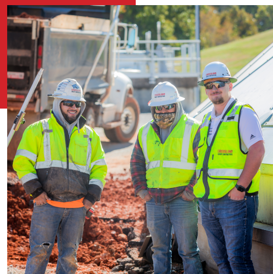 Three construction workers wearing hard hats, safety vests, and sunglasses stand together at a construction site with red dirt and a truck in the background on a sunny day.