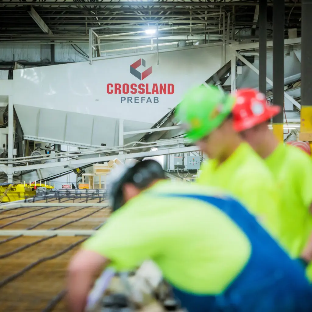 Three workers in safety gear focus on construction tasks inside a factory. The background shows industrial equipment and a large sign reading “Crossland Prefab.” The scene is brightly lit and emphasizes teamwork.