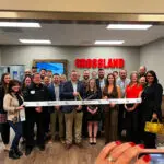 A group of people stand together smiling for a ribbon-cutting ceremony in an office lobby with a “CROSSLAND” sign on the wall behind them. The person in front holds large scissors and a ribbon with “Montrose” printed on it.