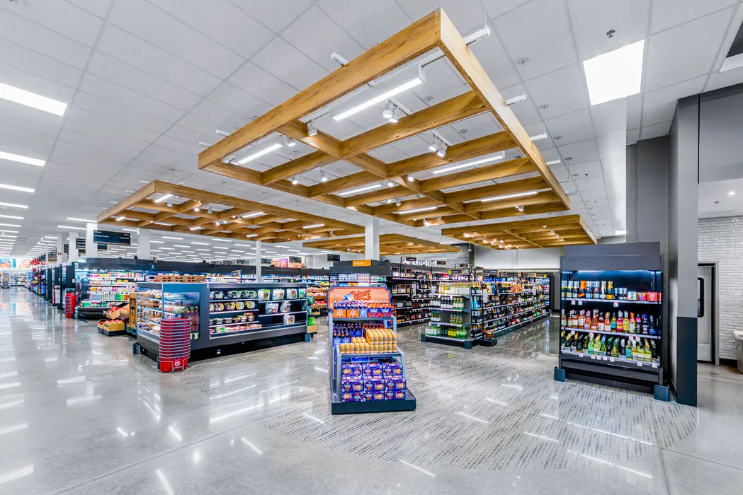 A brightly lit, modern grocery store interior with shiny floors, organized shelves of food and drinks, and a wooden ceiling feature above the aisles.